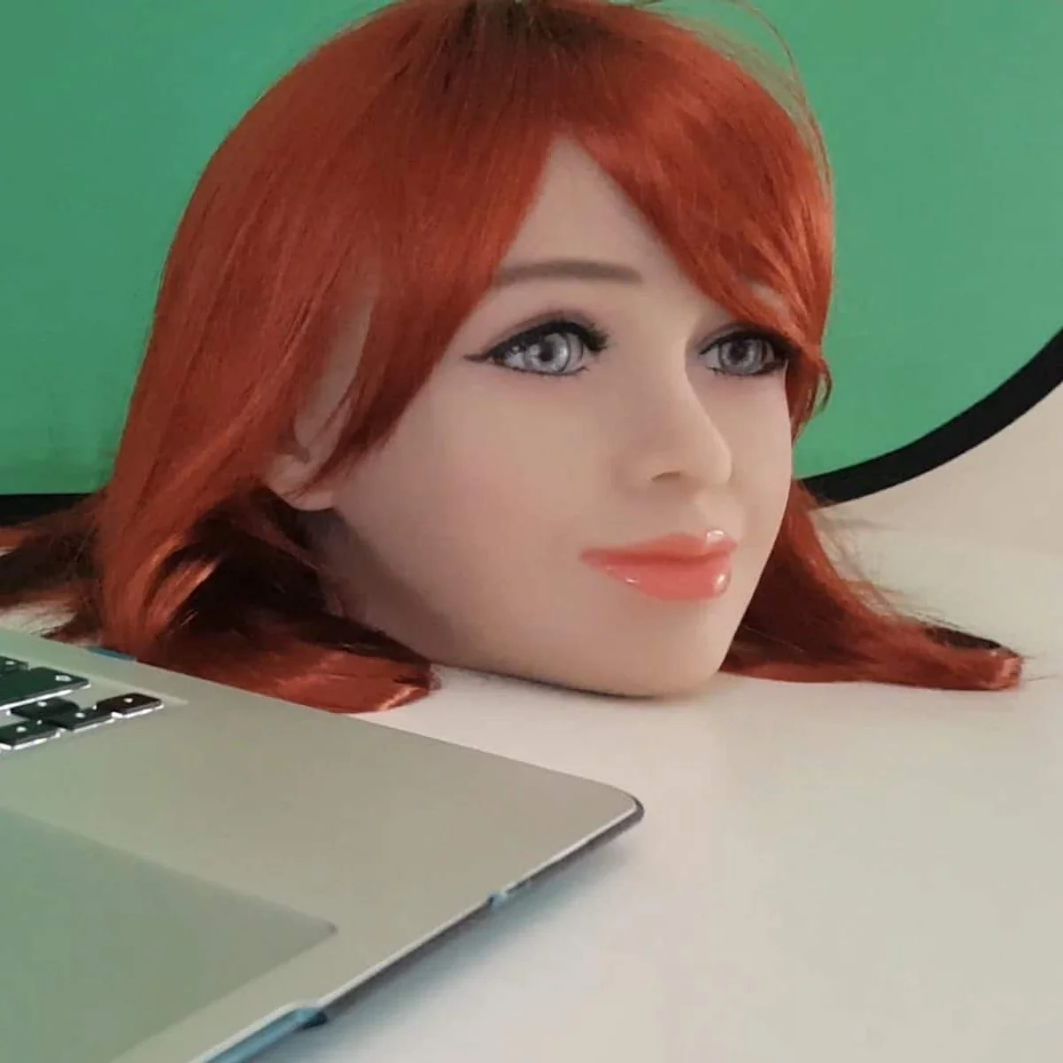 Et ældre Emma-robothoved med rød paryk fotograferet på et bord ved en bærbar computer